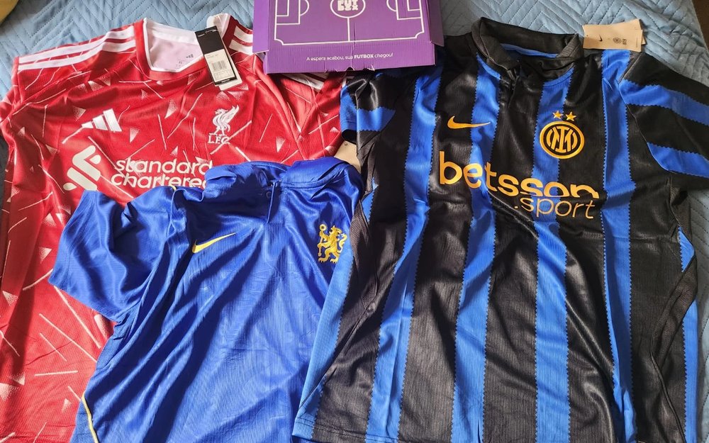 Caixa roxa da Futbox aberta com as camisas 26/27 do Liverpool, do Chelsea e da Inter de Milão, ainda com etiqueta
